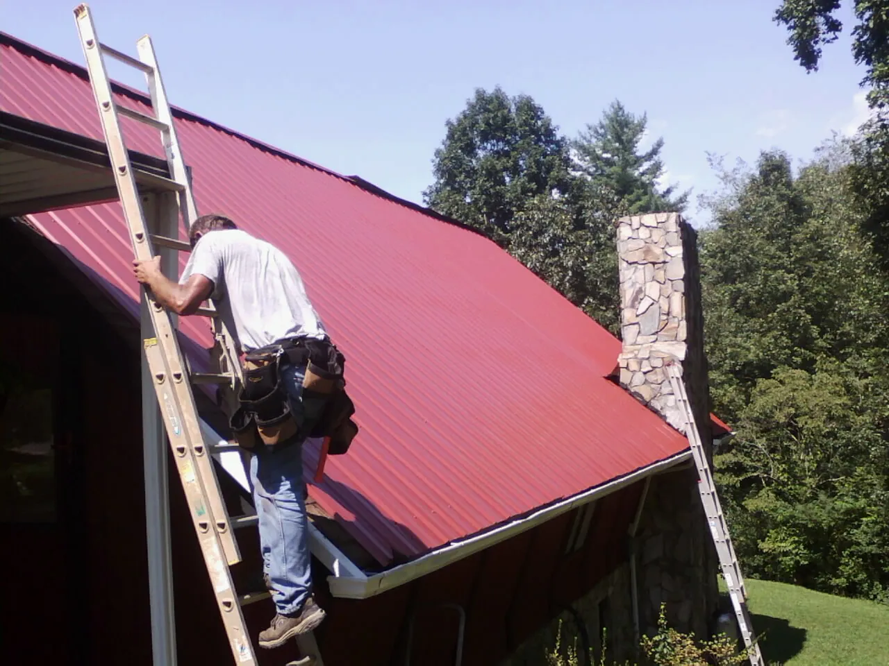 Roofer on a ladder beside a red metal roof and stone chimney — Soffit & Fascia Repair by Roofing Brothers in Uniondale